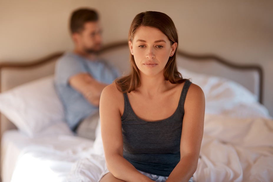 Woman sitting on edge of bed with man in background
